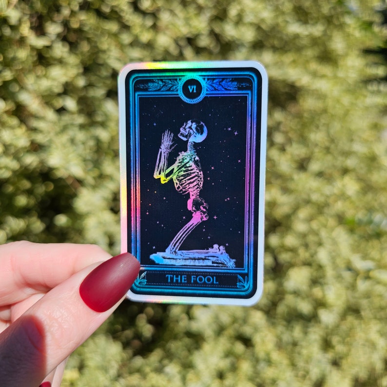May include: A holographic tarot card with a black background and a rainbow skeleton figure kneeling with hands raised. The card is labeled "The Fool" and has the Roman numeral "VI" in the top right corner.