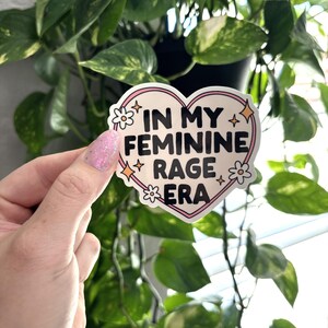 May include: A pink heart-shaped sticker with the text "In My Feminine Rage Era" in black lettering. The sticker has white flowers and glitter accents.