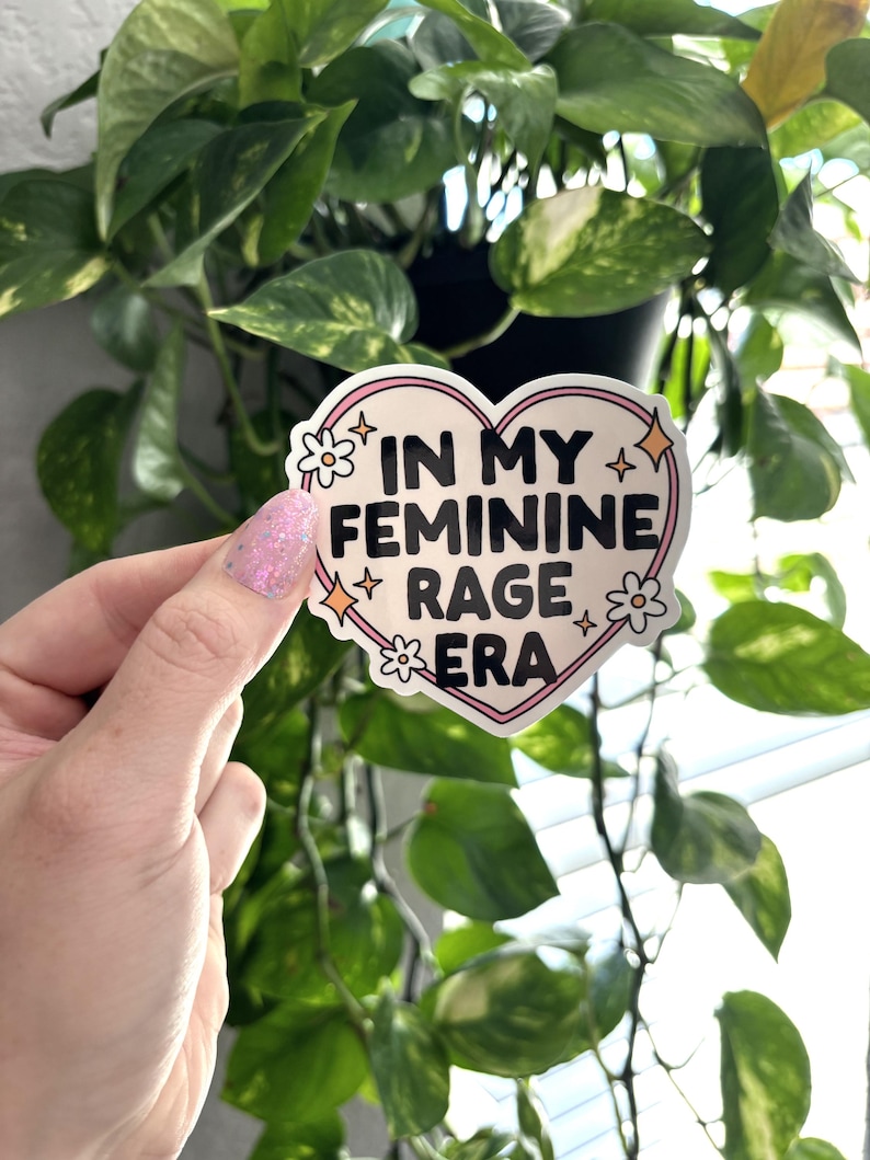 May include: A pink heart-shaped sticker with the text "In My Feminine Rage Era" in black lettering. The sticker has white flowers and glitter accents.