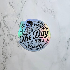May include: A holographic sticker with a skeleton holding up a peace sign and the text "Have the Day You Deserve."