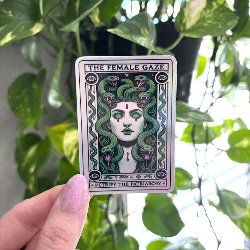 May include: A silver sticker with a green and white illustration of a woman with snakes for hair. The text "THE FEMALE GAZE" is at the top of the sticker and "PETRIFY THE PATRIARCHY" is at the bottom.