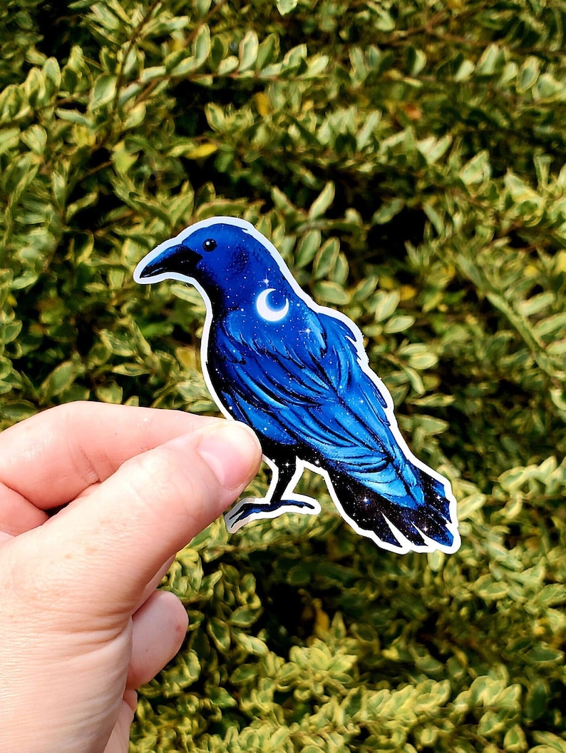 May include: A blue crow sticker with a crescent moon and stars on its feathers. The sticker is on a green leafy background.