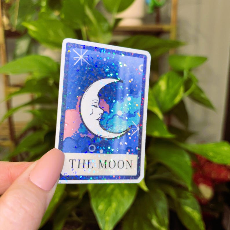 May include: A rectangular sticker featuring a crescent moon with a face, set against a blue, glittery background. The sticker has a white border and the words "THE MOON" at the bottom. The sticker is held in front of a green leafy plant.
