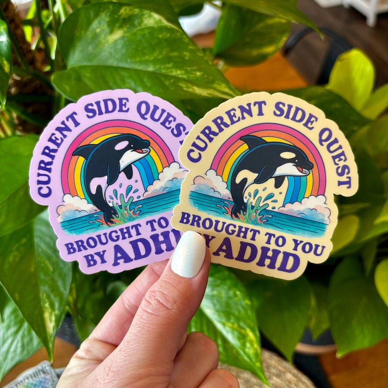 May include: Two stickers with a cartoon orca leaping from water under a rainbow. The stickers have the text "Current Side Quest" and "Brought to You by ADHD". One sticker is light purple, the other is light yellow.