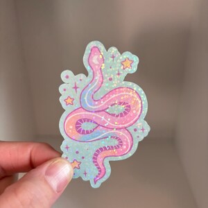 May include: A pink and purple iridescent glitter snake sticker with stars and a crescent moon on a light blue background.