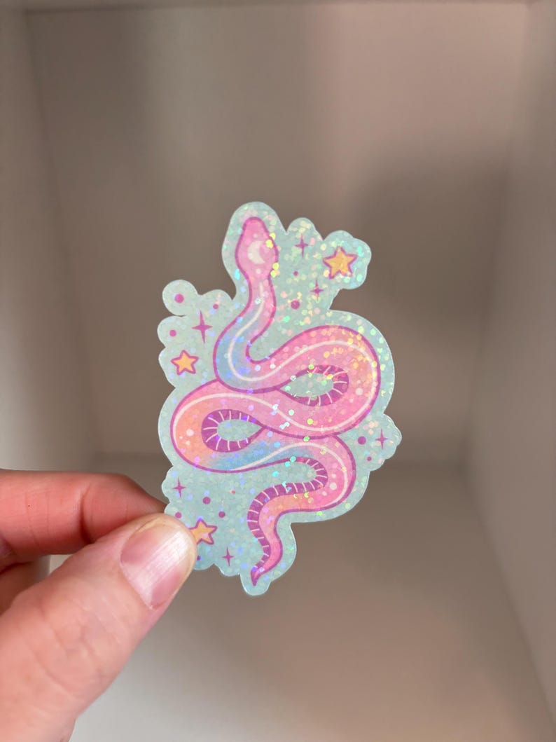 May include: A pink and purple iridescent glitter snake sticker with stars and a crescent moon on a light blue background.
