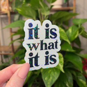 May include: A white sticker with iridescent glitter features the text "it is what it is" in black.