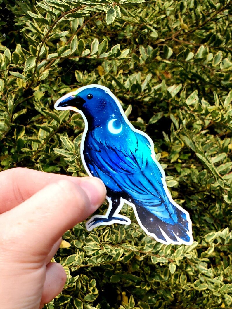 May include: A blue crow sticker with a crescent moon on its chest. The crow is depicted with a starry night sky pattern on its feathers.