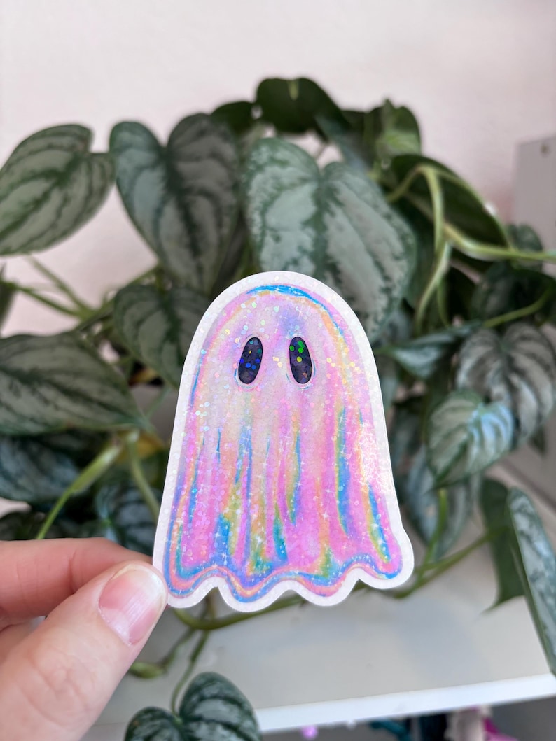 K&ouml;nnte beinhalten: Ein holografischer rosa-blauer Geisteraufkleber mit schwarzen Augen. Der Aufkleber befindet sich auf einem wei&szlig;en Hintergrund.