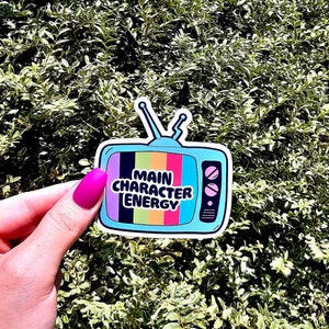 May include: A blue and white sticker featuring a retro television design. The television screen has a rainbow pattern and the text "MAIN CHARACTER ENERGY" in black.