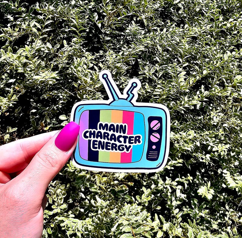 May include: A blue and white sticker featuring a retro television design. The television screen has a rainbow pattern and the text "MAIN CHARACTER ENERGY" in black.