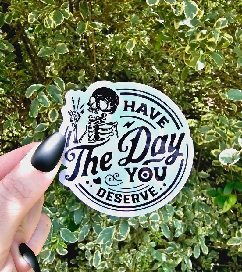 May include: A round sticker with a blue and white background. The sticker features a black skeleton with a peace sign and the text "Have the Day You Deserve."