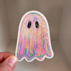 K&ouml;nnte beinhalten: Ein holografischer rosa und blauer Glitzer-Geisteraufkleber mit schwarzen Augen.