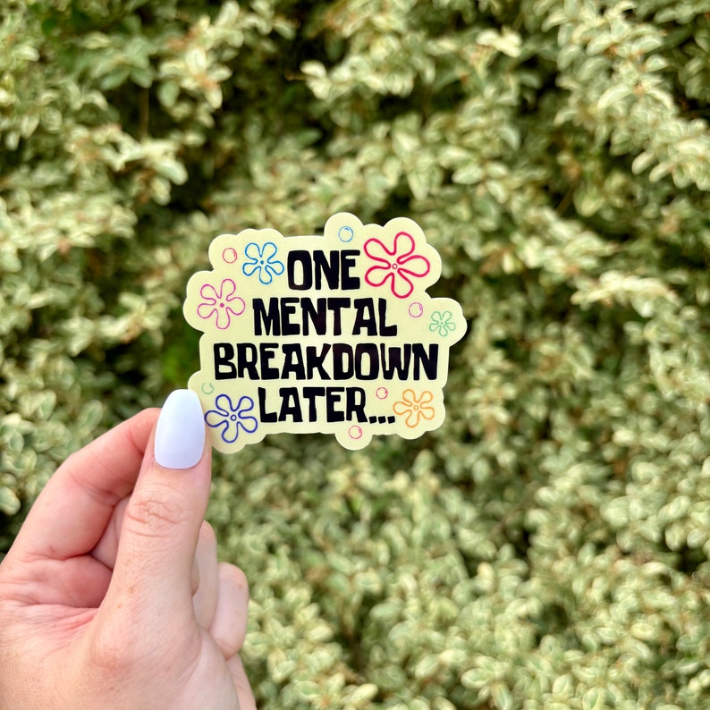 May include: A yellow sticker with the text "ONE MENTAL BREAKDOWN LATER..." in black. The sticker has a light green border and is decorated with colorful flower designs. The sticker is held by a hand with a light purple nail.
