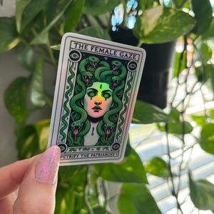 May include: A holographic sticker with a green and white illustration of a woman with snakes for hair. The text "THE FEMALE GAZE" and "PETRIFY THE PATRIARCHY" is printed on the sticker.