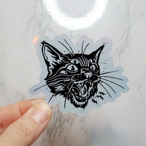 May include: Black and white sticker of a cat with multiple eyes and an open mouth, the cat is snarling with a menacing expression.