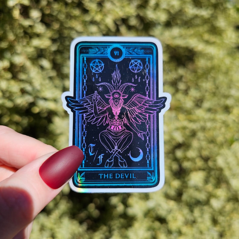 May include: A holographic tarot card sticker featuring the Devil card. The card is black with a pink and blue iridescent finish. The Devil is depicted as a goat-like figure with wings and a crown. The card also features the Roman numeral VI and the text "THE DEVIL".