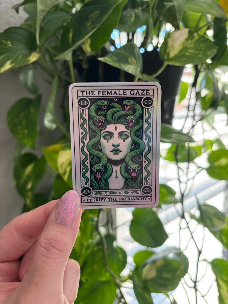 May include: A silver sticker with a green and black illustration of a woman with snakes for hair. The text "THE FEMALE GAZE" is at the top of the sticker and "PETRIFY THE PATRIARCHY" is at the bottom.
