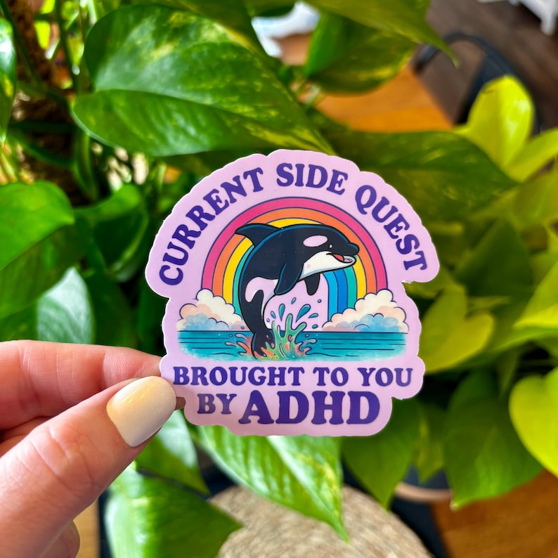 May include: A sticker with the text "CURRENT SIDE QUEST" and "BROUGHT TO YOU BY ADHD". The sticker features a cartoon orca leaping through a rainbow over ocean waves. The sticker is held by a hand.