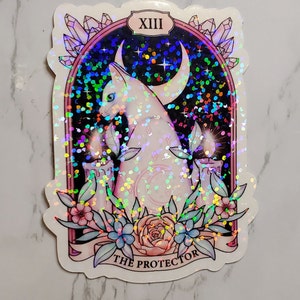 May include: A holographic sticker featuring a white cat with a crescent moon behind it. The cat is sitting in front of two lit candles and surrounded by flowers. The sticker has a pink border and the text "XIII THE PROTECTOR" at the bottom.