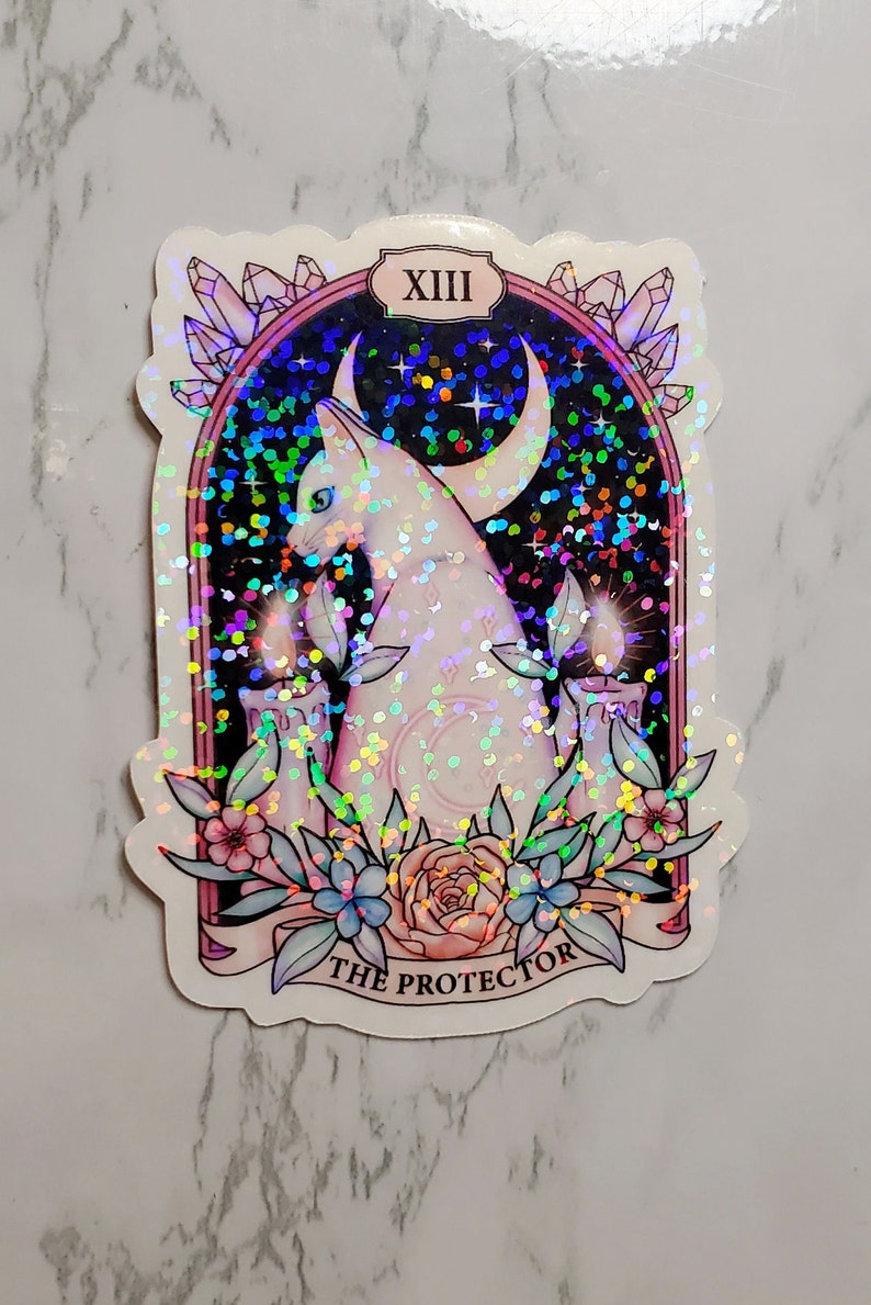 May include: A holographic sticker featuring a white cat with a crescent moon behind it. The cat is sitting in front of two lit candles and surrounded by flowers. The sticker has a pink border and the text "XIII THE PROTECTOR" at the bottom.