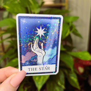 May include: A tarot card with a blue background and a hand reaching up to a star. The card is decorated with glitter and the text "THE STAR" is printed at the bottom.