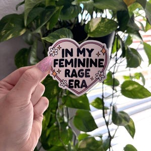 May include: A pink heart-shaped sticker with the text "In My Feminine Rage Era" in black lettering. The sticker has a white outline and small white flowers and stars around the text.