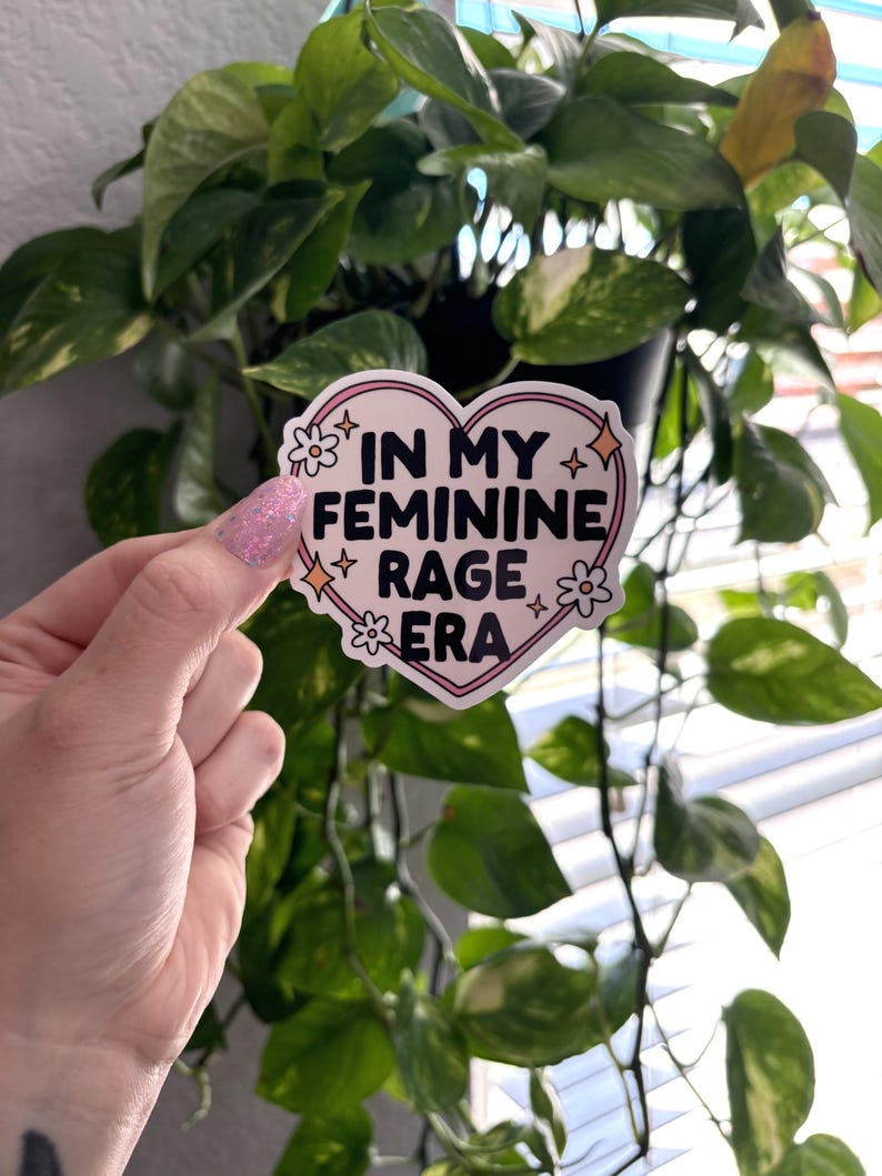 May include: A pink heart-shaped sticker with the text "In My Feminine Rage Era" in black lettering. The sticker has a white outline and small white flowers and stars around the text.