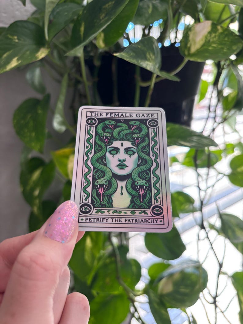 May include: A sticker with a holographic finish featuring a green-haired woman with snakes around her head. The text on the sticker reads "The Female Gaze" and "Petrify the Patriarchy".
