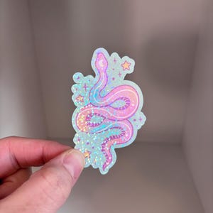 May include: A holographic sticker featuring a pink and blue snake with a glittery finish. The snake is curled up with its tail in its mouth. There are small stars around the snake.