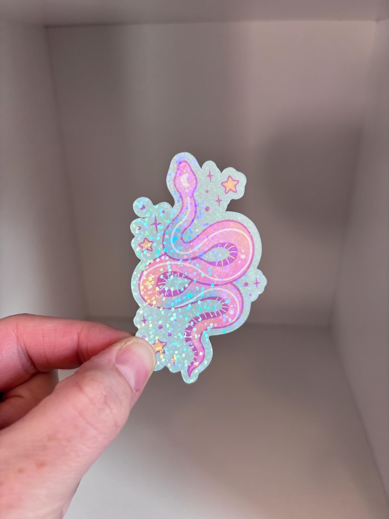 May include: A holographic sticker featuring a pink and blue snake with a glittery finish. The snake is curled up with its tail in its mouth. There are small stars around the snake.