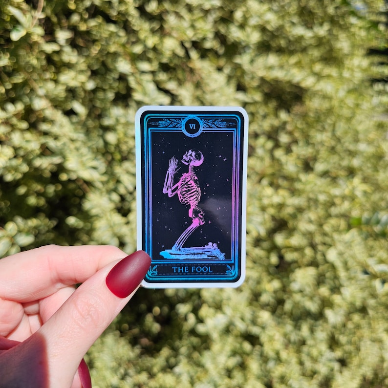 May include: A tarot card with a holographic finish depicting a skeleton in a kneeling pose. The card is labeled "The Fool" and has the Roman numeral "VI" in the top right corner.