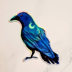 May include: A blue iridescent sticker of a crow with a crescent moon and stars on its chest. The crow is standing on a white background.