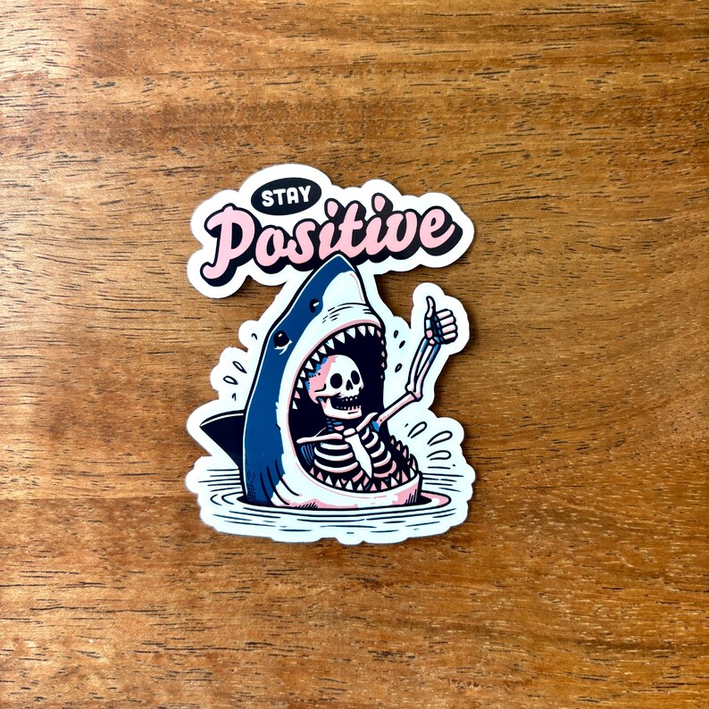 May include: A sticker featuring a cartoon shark with a skeleton inside, giving a thumbs-up. The shark is blue and white, with the skeleton in white. The sticker has the words "Stay Positive" in pink and white.