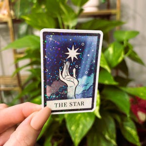May include: A holographic sticker featuring The Star tarot card. The card depicts a hand reaching up to a star in a blue and purple sky with white clouds. The card has a glittery finish.