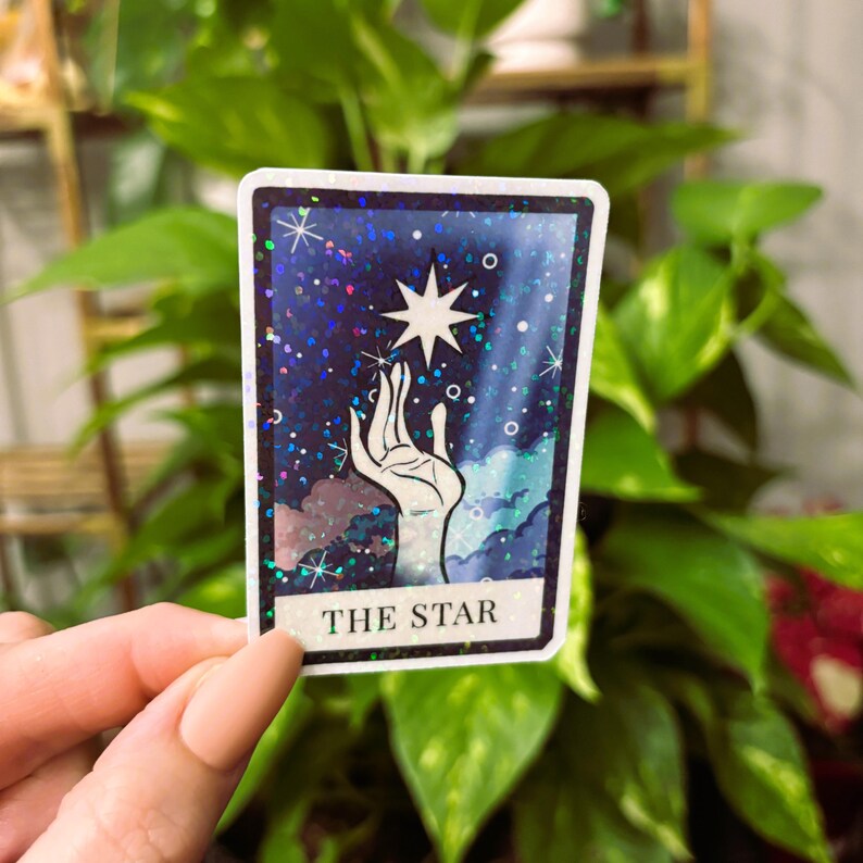 May include: A holographic sticker featuring The Star tarot card. The card depicts a hand reaching up to a star in a blue and purple sky with white clouds. The card has a glittery finish.