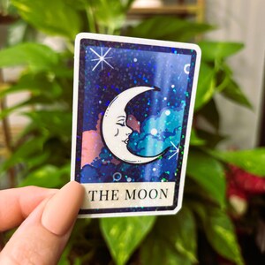 May include: A tarot card sticker featuring a crescent moon with a face against a starry, blue background. The card is labeled "THE MOON" in white text at the bottom. The sticker has a white border and is held in front of a green leafy background.