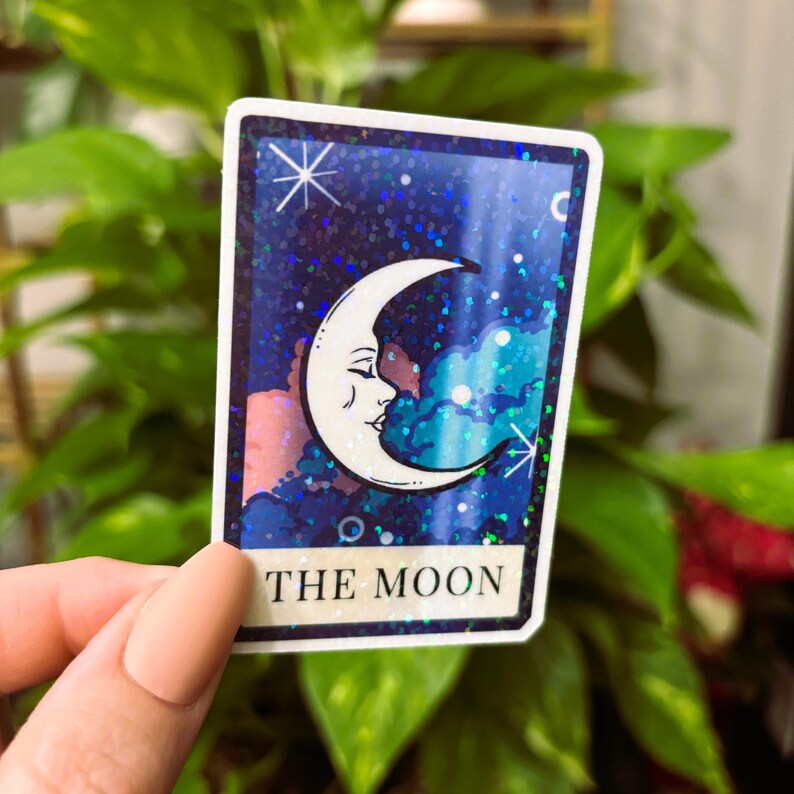 May include: A tarot card sticker featuring a crescent moon with a face against a starry, blue background. The card is labeled "THE MOON" in white text at the bottom. The sticker has a white border and is held in front of a green leafy background.