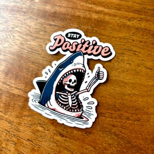 May include: A sticker featuring a cartoon shark with an open mouth, revealing a skeleton giving a thumbs-up. The sticker has the words "Stay Positive" in a pink and black font.