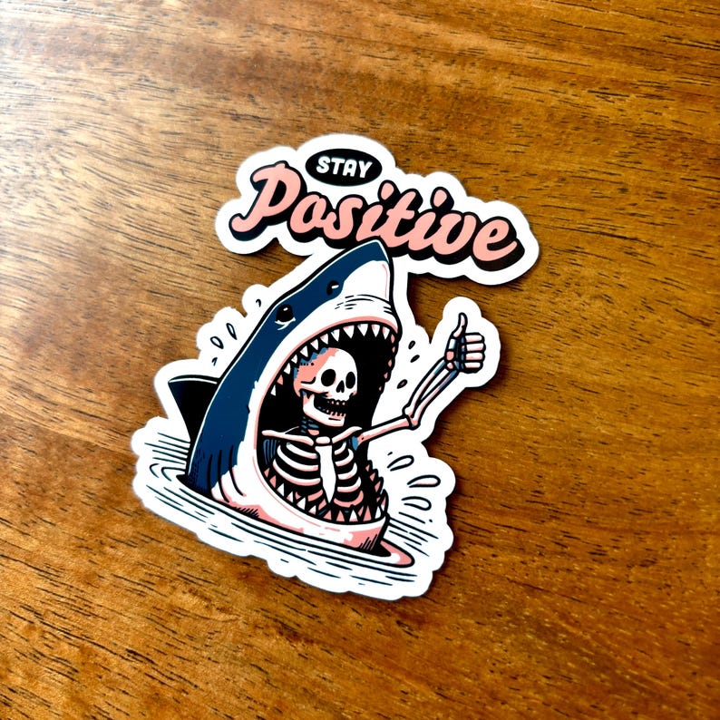 May include: A sticker featuring a cartoon shark with an open mouth, revealing a skeleton giving a thumbs-up. The sticker has the words "Stay Positive" in a pink and black font.