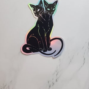 May include: A black and white sticker of a two-headed cat sitting with its tail curled around its body. The sticker has a holographic finish.