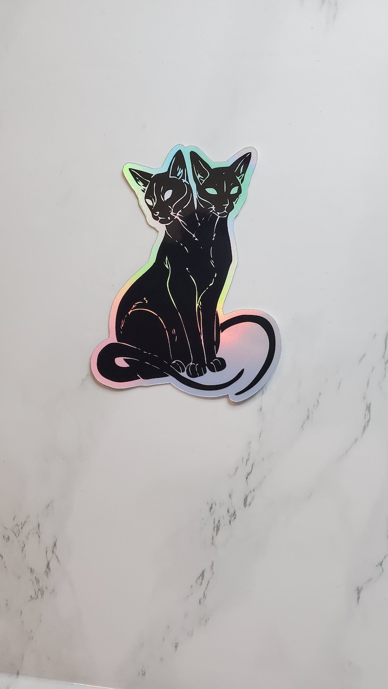 May include: A black and white sticker of a two-headed cat sitting with its tail curled around its body. The sticker has a holographic finish.