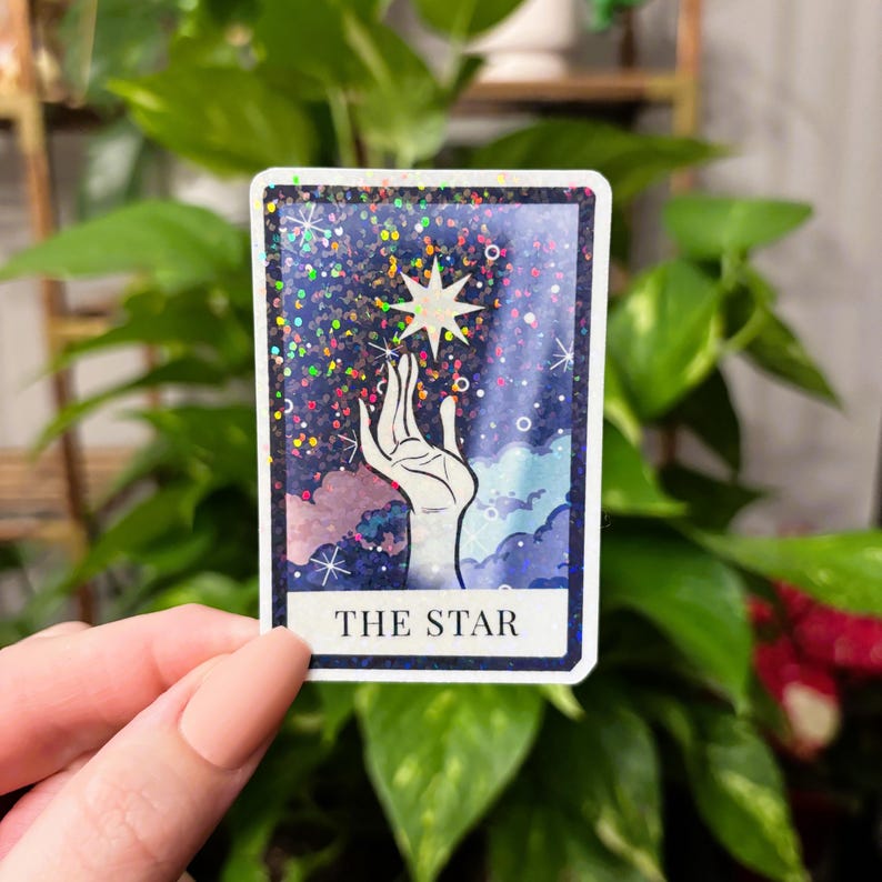 May include: A holographic sticker featuring The Star tarot card. The card depicts a hand reaching up towards a star in a night sky with clouds.