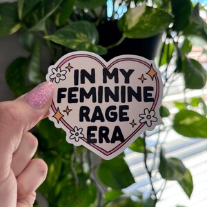 May include: A pink heart-shaped sticker with the text "In My Feminine Rage Era" in black lettering. The sticker is decorated with white flowers and gold glitter.