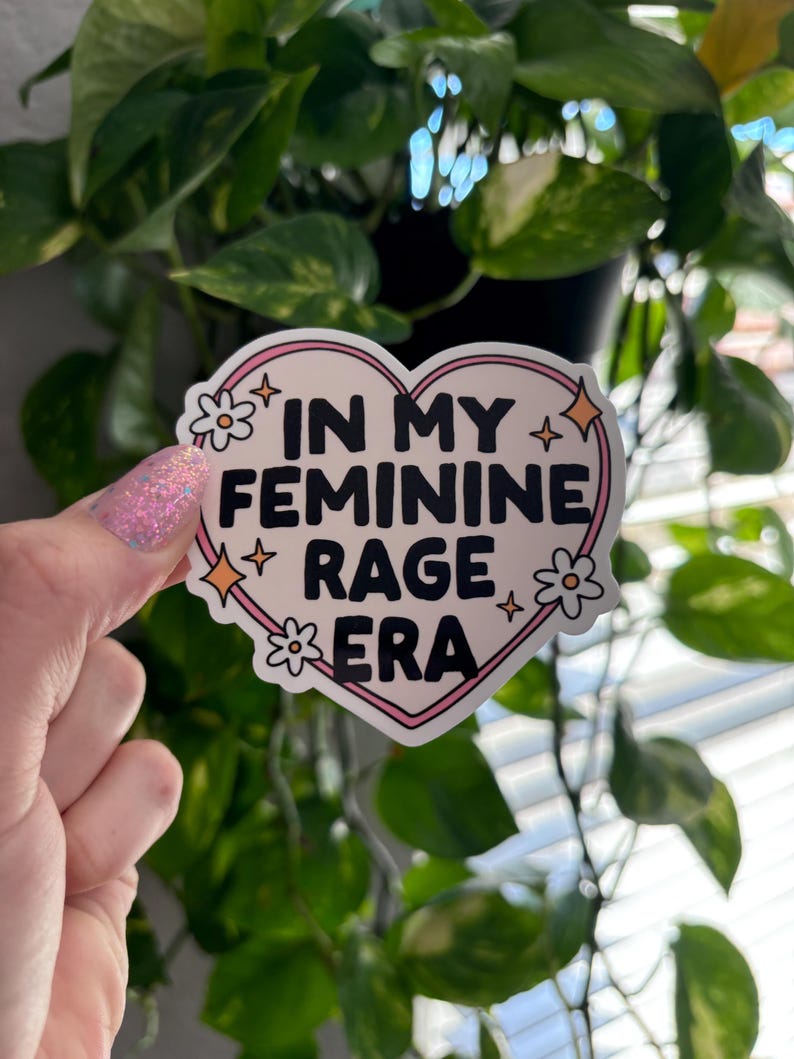 May include: A pink heart-shaped sticker with the text "In My Feminine Rage Era" in black lettering. The sticker is decorated with white flowers and gold glitter.
