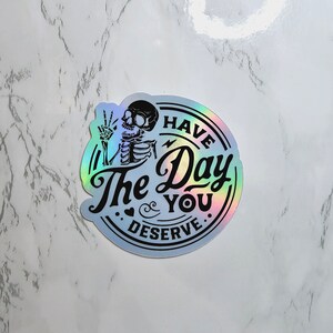 May include: A holographic sticker with a skeleton holding up a peace sign and the text "Have the Day You Deserve."