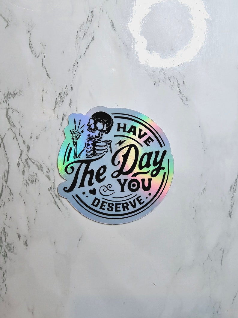 May include: A holographic sticker with a skeleton holding up a peace sign and the text "Have the Day You Deserve."