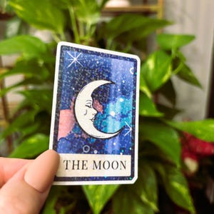 May include: A rectangular sticker featuring a crescent moon with a face against a starry night sky. The card reads "THE MOON" in white text at the bottom. The background is a mix of blue, pink, and purple with glitter accents.