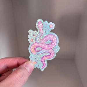 May include: A holographic sticker featuring a pink and blue snake with stars on a light blue background.