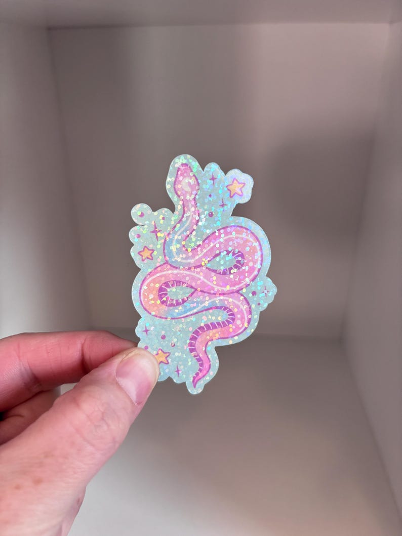 May include: A holographic sticker featuring a pink and blue snake with stars on a light blue background.